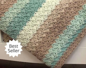 May include: A crocheted baby blanket with a chevron pattern in shades of beige, white, and teal. The blanket is draped over a white crib and has a "Best Seller" tag attached.