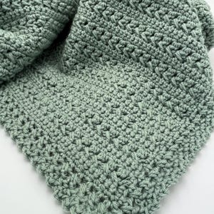 May include: A soft, sage green crocheted blanket with a delicate stitch pattern. The blanket is folded in half, showing the texture of the fabric.