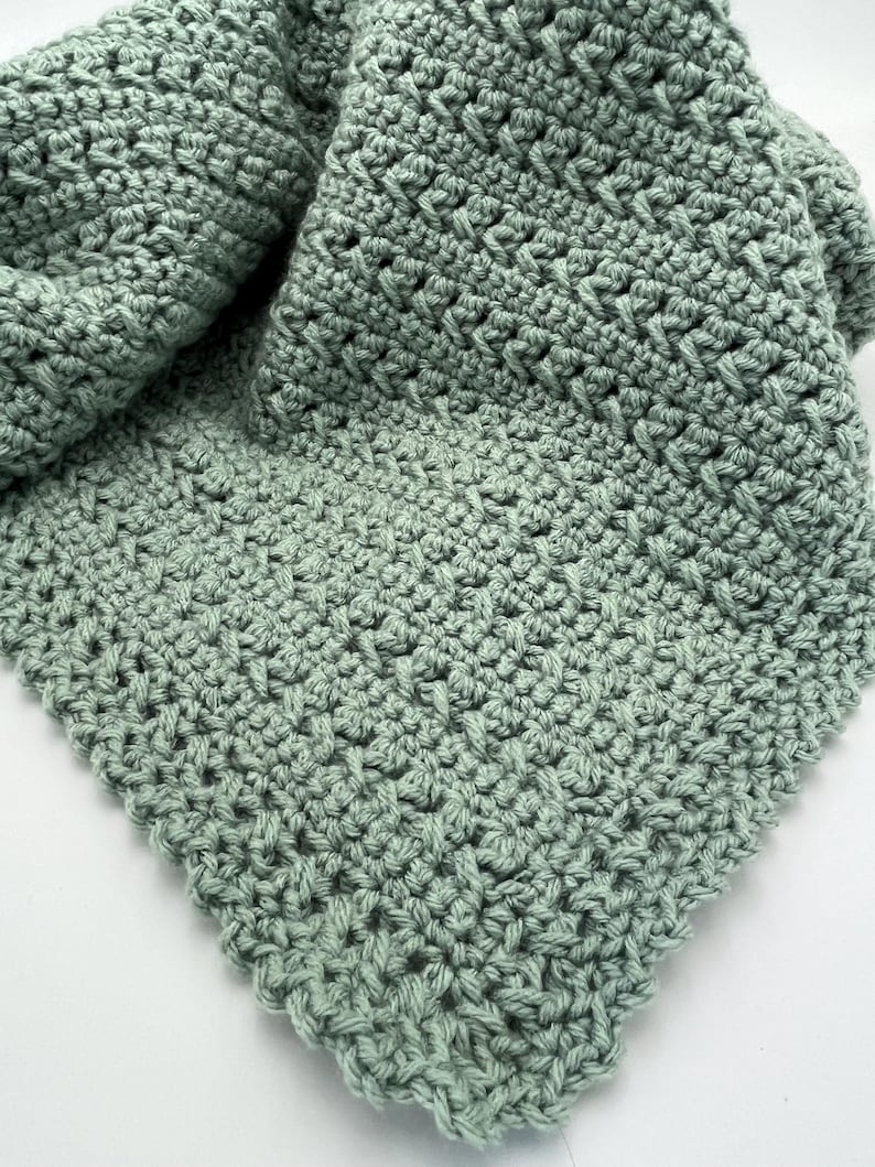 May include: A soft, sage green crocheted blanket with a delicate stitch pattern. The blanket is folded in half, showing the texture of the fabric.