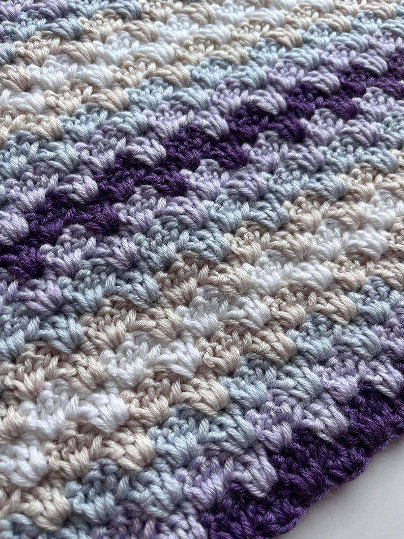 May include: A close-up of a crocheted blanket with stripes of purple, light blue, white, and beige yarn.