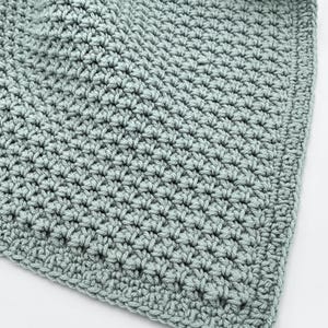 May include: Close-up of a soft, crocheted blanket in a muted teal color. The blanket features a textured stitch pattern, with a visible border. The handmade blanket is likely made of a soft yarn, perfect for a baby or as a decorative throw.