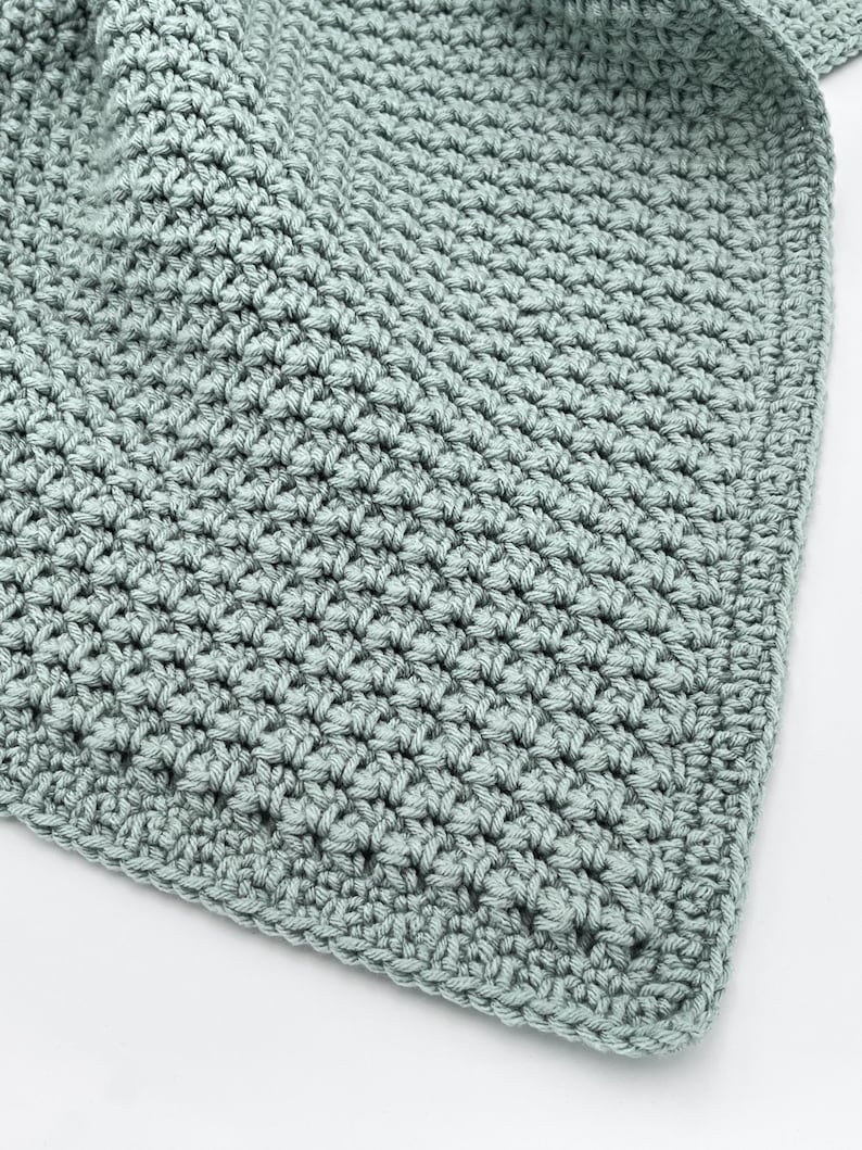 May include: Close-up of a soft, crocheted blanket in a muted teal color. The blanket features a textured stitch pattern, with a visible border. The handmade blanket is likely made of a soft yarn, perfect for a baby or as a decorative throw.