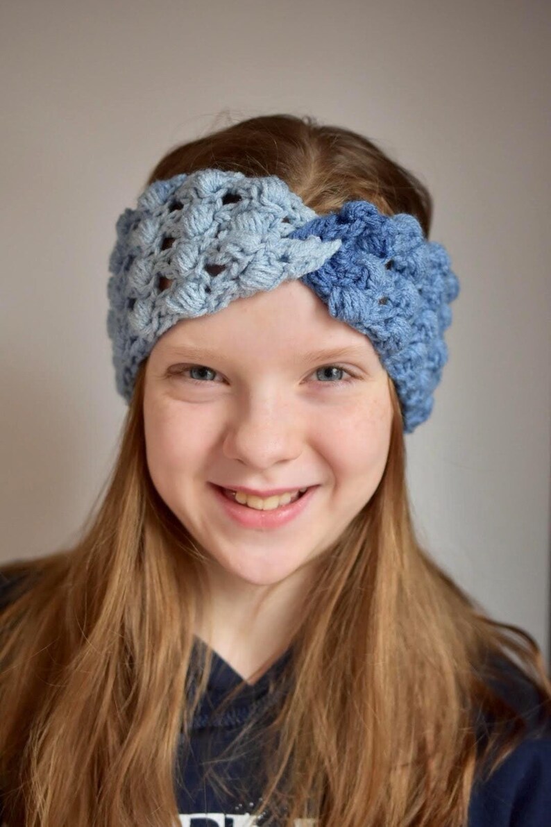 May include: A young person with long brown hair wearing a blue and light blue crocheted headband. The headband is a twisted design.