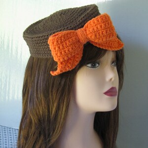 May include: A brown crocheted hat with a large orange crocheted bow. The hat is worn on a mannequin with brown hair.