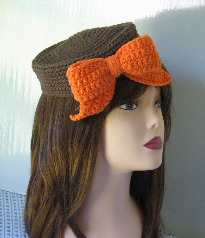 May include: A brown crocheted hat with a large orange crocheted bow. The hat is worn on a mannequin with brown hair.