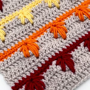 Puede incluir: Primer plano de un artículo de ganchillo con fondo gris. Filas diagonales de diseños de hojas amarillas, naranjas y rojas están cosidas sobre la tela gris. Las hojas son de un color contrastante con las filas naranja y amarilla.