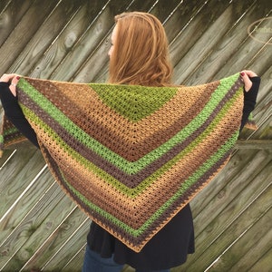 Könnte beinhalten: Ein gehäkeltes Tuch in Grün-, Braun- und Beigetönen. Das Tuch ist dreieckig und hat ein gestreiftes Muster. Das Tuch wird von einer Person mit langen roten Haaren getragen.