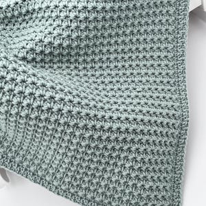 May include: A close-up of a soft, textured, light teal crocheted blanket. The blanket has a tight stitch pattern, creating a dense, warm appearance. The edges are neatly finished, suggesting a handmade item, perfect for home decor.