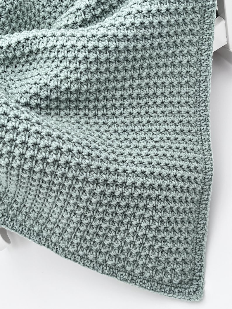 May include: A close-up of a soft, textured, light teal crocheted blanket. The blanket has a tight stitch pattern, creating a dense, warm appearance. The edges are neatly finished, suggesting a handmade item, perfect for home decor.