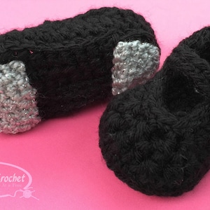 Puede incluir: Dos zapatos de bebé de ganchillo negros con detalles plateados. Un zapato tiene un cierre de hebilla. Los zapatos están sobre una superficie rosa.