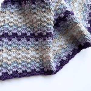May include: A crocheted blanket with a purple border and a gradient of light blue, beige, and purple colors.