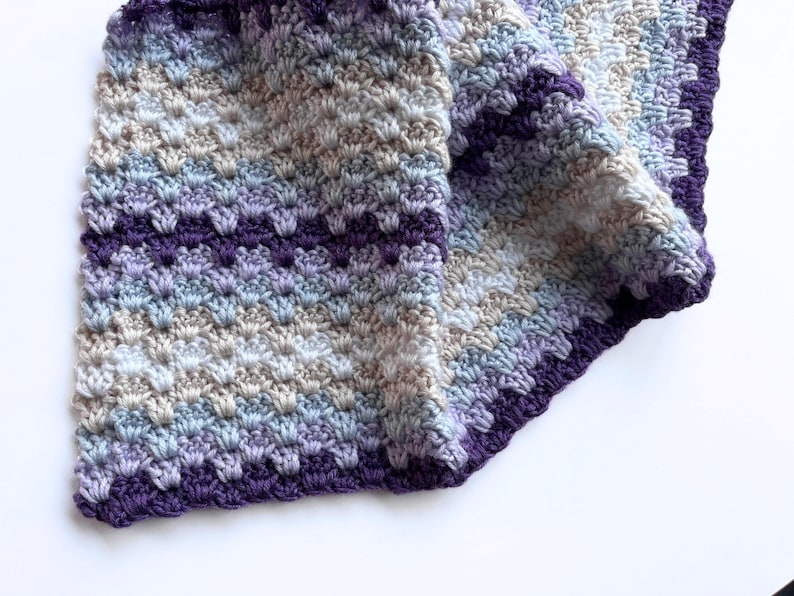 May include: A crocheted blanket with a purple border and a gradient of light blue, beige, and purple colors.