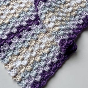 May include: A close-up of a crocheted blanket with a purple, white, and gray color scheme. The blanket is made with a textured stitch that creates a bumpy surface.
