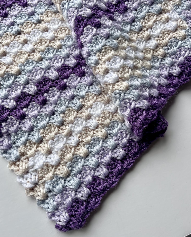 May include: A close-up of a crocheted blanket with a purple, white, and gray color scheme. The blanket is made with a textured stitch that creates a bumpy surface.