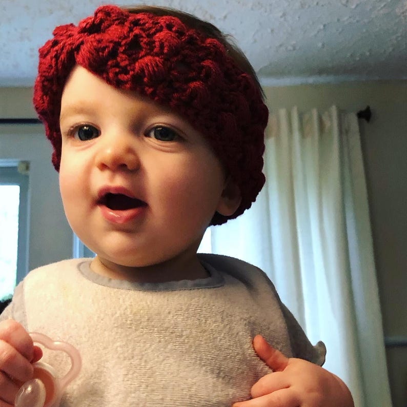 May include: A young child wearing a red crocheted headband. The headband is a twisted design and covers the child's forehead.