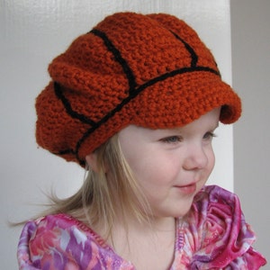 May include: A young child wearing a crocheted orange and black basketball-themed hat with a brim. The hat has a black stitching design that resembles a basketball.