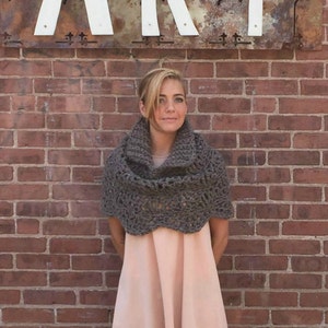Puede incluir: Una mujer lleva una bufanda de cuello de tortuga de ganchillo marrón con un borde festoneado. Está de pie frente a una pared de ladrillo.