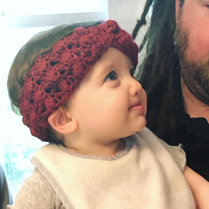 May include: A close-up of a baby wearing a burgundy crocheted headband. The baby is looking up and smiling.