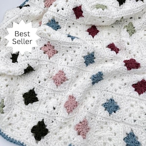 May include: A white crocheted blanket with a floral pattern in various colors, including pink, blue, and burgundy. The blanket has a scalloped edge with a blue trim. The text "Best Seller" is in a white starburst.