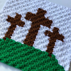 May include: A handmade crochet piece featuring three brown crosses against a white background, with a green grassy area at the bottom. The textured stitches create a three-dimensional effect, showcasing the detailed craftsmanship of the religious-themed artwork.