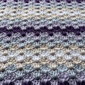 May include: A close-up of a crocheted blanket with a chevron pattern in shades of purple, blue, white, and beige.