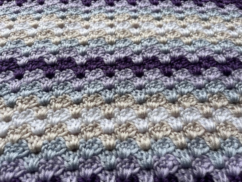 May include: A close-up of a crocheted blanket with a chevron pattern in shades of purple, blue, white, and beige.