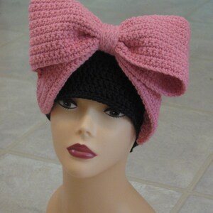 May include: A pink crocheted bow sits atop a black crocheted beanie. The beanie is worn on a mannequin head.