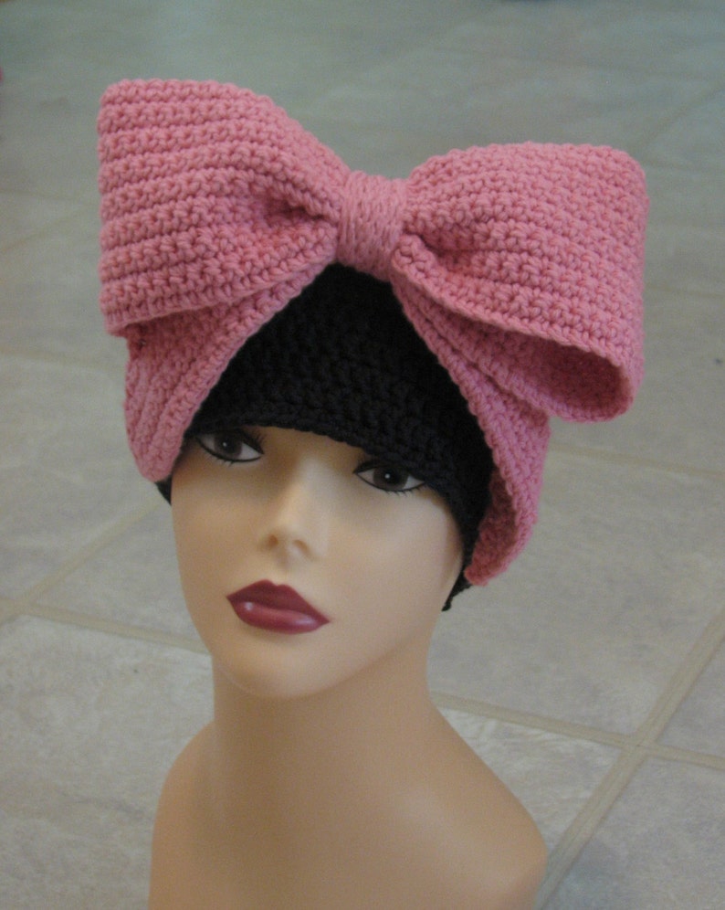 May include: A pink crocheted bow sits atop a black crocheted beanie. The beanie is worn on a mannequin head.