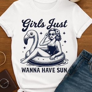 May include: White t-shirt with a graphic of a woman in a swimsuit on a flamingo float. The text "Girls Just Wanna Have Sun" is above and below the image in navy blue. The shirt is on a wooden surface.