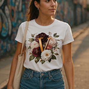Puede incluir: Camiseta blanca con diseño floral y la letra dorada "V". Las flores son burdeos, blancas y rosas, con hojas verdes. La modelo lleva shorts vaqueros.