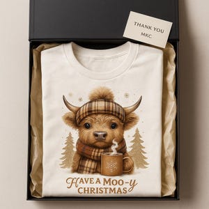 May include: A cream-colored t-shirt featuring a Highland cow wearing a plaid hat and scarf, holding a mug with steam. The shirt is inside a black gift box with a card that says "Thank You MKC". The text on the shirt says "Have a Moo-y Christmas".