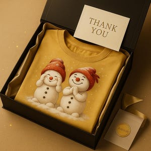 May include: A yellow t-shirt featuring two snowmen with red hats and carrot noses, inside a black gift box. The snowmen are smiling and have black button eyes. A card with "THANK YOU" is on top. A small gift bag with a gold seal is also included.