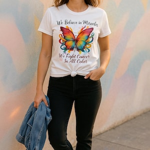May include: White t-shirt with a colorful butterfly graphic and the text "We Believe in Miracles" and "We Fight Cancer In All Color." The person is wearing black jeans and white sneakers, holding a blue denim jacket.