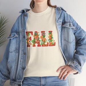 White t-shirt folded in a gift box with orange tissue paper, featuring the word “FALL” in floral lettering with leaves and flowers. Decorated with a brown tag reading “Happy Fall,” plaid ribbon, and acorns on a wooden table. Cozy rustic autumn aesthetic perfect for fall gifts and seasonal outfits.