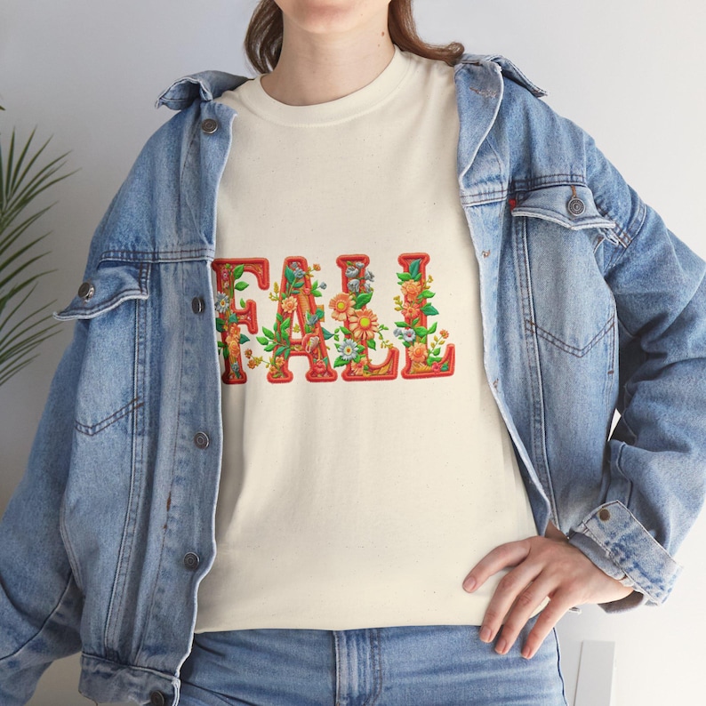 White t-shirt folded in a gift box with orange tissue paper, featuring the word “FALL” in floral lettering with leaves and flowers. Decorated with a brown tag reading “Happy Fall,” plaid ribbon, and acorns on a wooden table. Cozy rustic autumn aesthetic perfect for fall gifts and seasonal outfits.