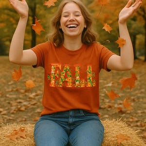 White t-shirt folded in a gift box with orange tissue paper, featuring the word “FALL” in floral lettering with leaves and flowers. Decorated with a brown tag reading “Happy Fall,” plaid ribbon, and acorns on a wooden table. Cozy rustic autumn aesthetic perfect for fall gifts and seasonal outfits.