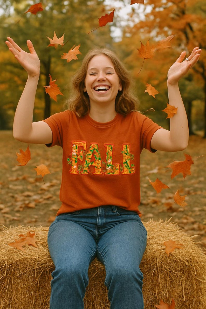 White t-shirt folded in a gift box with orange tissue paper, featuring the word “FALL” in floral lettering with leaves and flowers. Decorated with a brown tag reading “Happy Fall,” plaid ribbon, and acorns on a wooden table. Cozy rustic autumn aesthetic perfect for fall gifts and seasonal outfits.