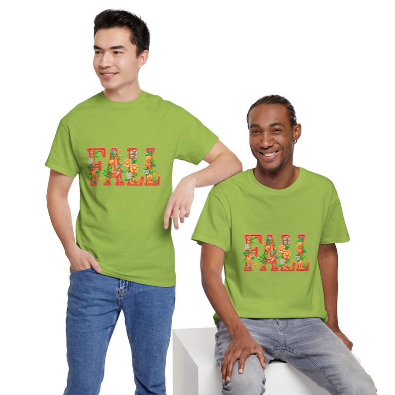 White t-shirt folded in a gift box with orange tissue paper, featuring the word “FALL” in floral lettering with leaves and flowers. Decorated with a brown tag reading “Happy Fall,” plaid ribbon, and acorns on a wooden table. Cozy rustic autumn aesthetic perfect for fall gifts and seasonal outfits.