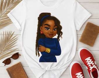 May include: White t-shirt featuring a cartoon illustration of a young Black woman with long brown hair, wearing a blue top and gold bracelets. The illustration has a smiling expression. The shirt is on a wooden surface.