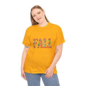 White t-shirt folded in a gift box with orange tissue paper, featuring the word “FALL” in floral lettering with leaves and flowers. Decorated with a brown tag reading “Happy Fall,” plaid ribbon, and acorns on a wooden table. Cozy rustic autumn aesthetic perfect for fall gifts and seasonal outfits.