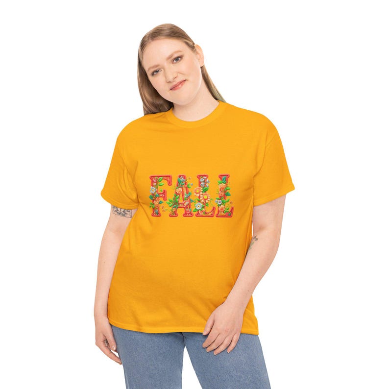 White t-shirt folded in a gift box with orange tissue paper, featuring the word “FALL” in floral lettering with leaves and flowers. Decorated with a brown tag reading “Happy Fall,” plaid ribbon, and acorns on a wooden table. Cozy rustic autumn aesthetic perfect for fall gifts and seasonal outfits.