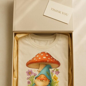 Könnte beinhalten: Ein cremefarbenes T-Shirt mit einer skurrilen Illustration eines Zwergs, der unter einem Pilz mit roter und weiß gepunkteter Kappe sitzt. Das Shirt befindet sich in einer offenen Geschenkbox mit einer "THANK YOU"-Karte.