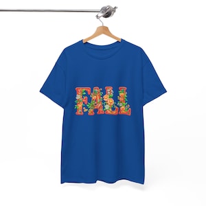 White t-shirt folded in a gift box with orange tissue paper, featuring the word “FALL” in floral lettering with leaves and flowers. Decorated with a brown tag reading “Happy Fall,” plaid ribbon, and acorns on a wooden table. Cozy rustic autumn aesthetic perfect for fall gifts and seasonal outfits.