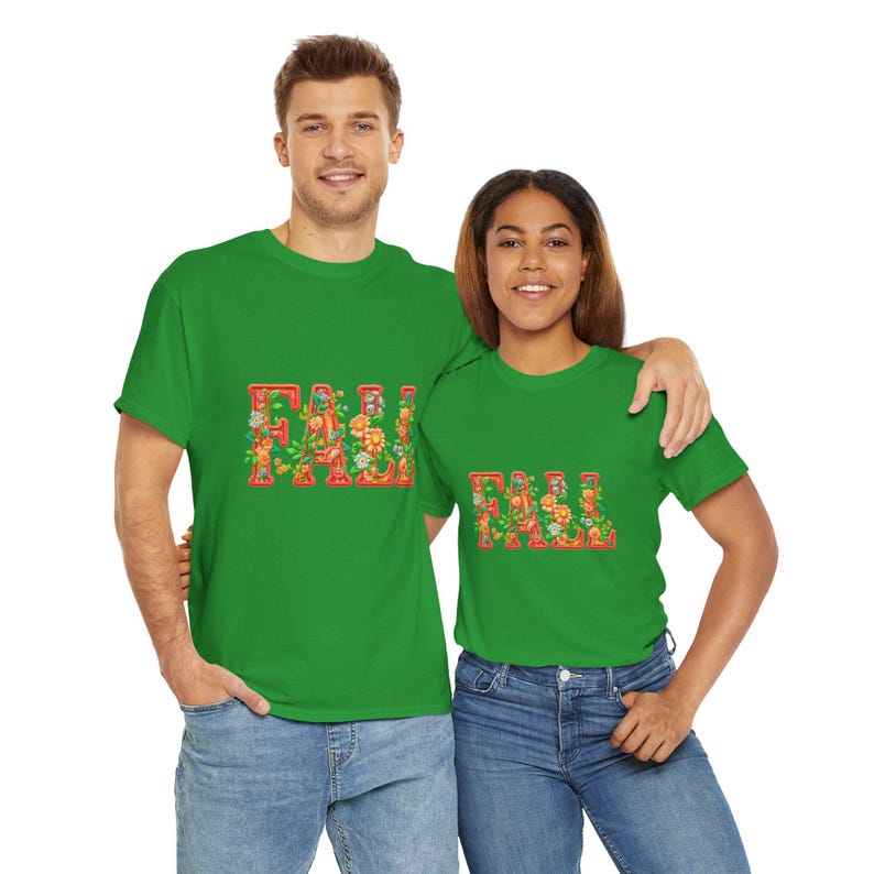 White t-shirt folded in a gift box with orange tissue paper, featuring the word “FALL” in floral lettering with leaves and flowers. Decorated with a brown tag reading “Happy Fall,” plaid ribbon, and acorns on a wooden table. Cozy rustic autumn aesthetic perfect for fall gifts and seasonal outfits.