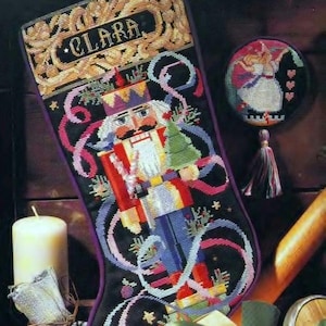 May include: A black Christmas stocking with a cross-stitch design featuring a nutcracker, ribbons, and the name "CLARA." A round ornament with a ballerina and a tassel hangs nearby. A lit candle and wrapped gift are also present.