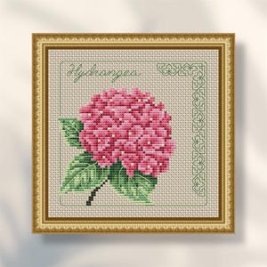 May include: Cross-stitch embroidery of a pink hydrangea flower with green leaves and stem, framed in a gold-colored frame. The word "Hydrangea" is stitched in green above the flower.