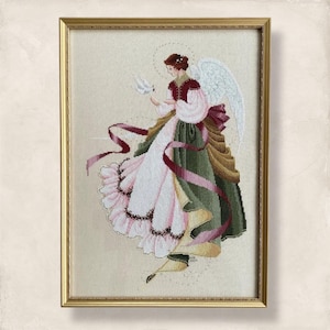 May include: Cross-stitch artwork of an angel in a gold frame. The angel wears a white dress with pink and green accents, holding a white dove. Long pink ribbons flow from the angel's waist. The angel has white wings and a halo.