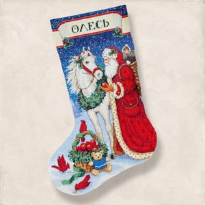Puede incluir: Calcetín navideño bordado con Papá Noel, un caballo blanco con una corona y una cesta de manzanas. El calcetín tiene un fondo azul con nieve y el nombre "Олесь". También se presentan pájaros rojos y un oso de peluche.
