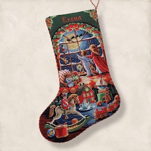 May include: A festive Christmas stocking with a detailed cross-stitch design. The stocking depicts a winter scene with children, toys, and a snowy night sky. The name "Elena" is embroidered at the top. The color palette includes red, green, blue, and brown.