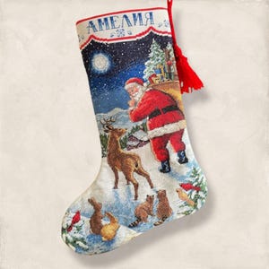 Puede incluir: Calcetín navideño bordado con una escena invernal que presenta a Papá Noel, renos y animales del bosque. El calcetín es blanco con una borla roja y el nombre "АМЕЛИЯ" bordado en la parte superior. El diseño incluye un paisaje nevado con luna llena.